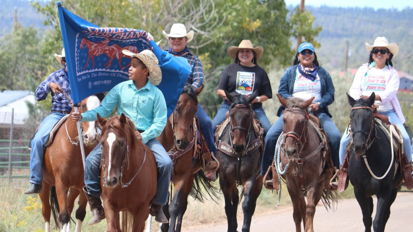 A group of Native American riders on horseback participate in a voter mobilization event, with a young rider at the front holding a blue flag that reads, 