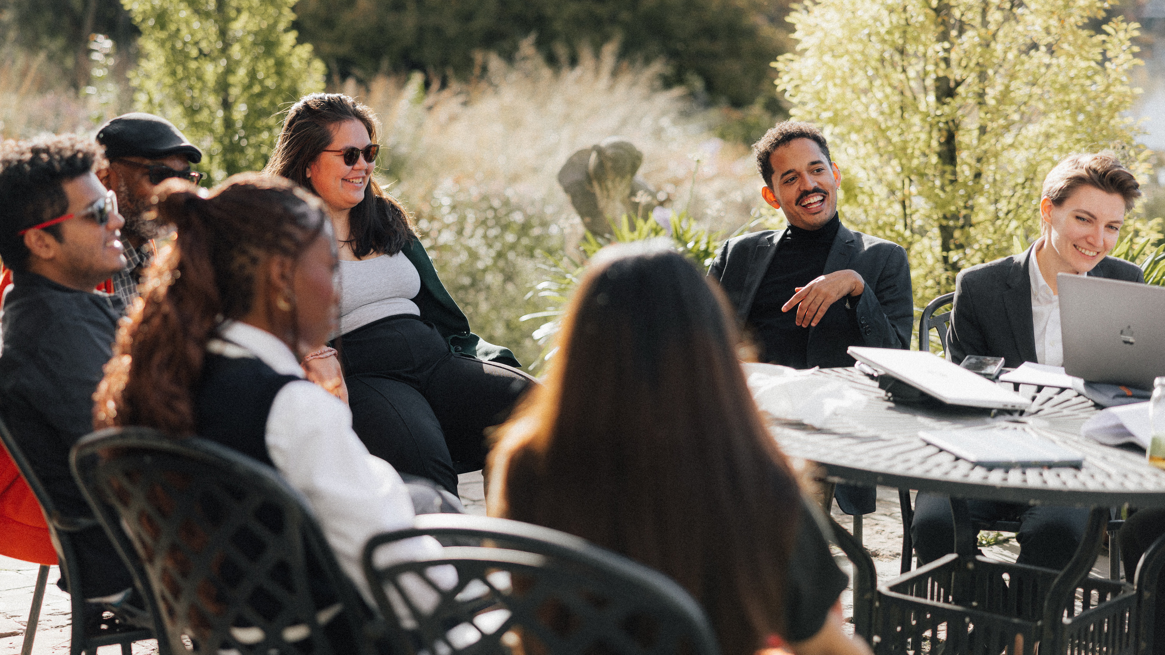 A diverse group of people sitting outdoors around a table, engaging in a lively discussion with smiles and positive expressions.