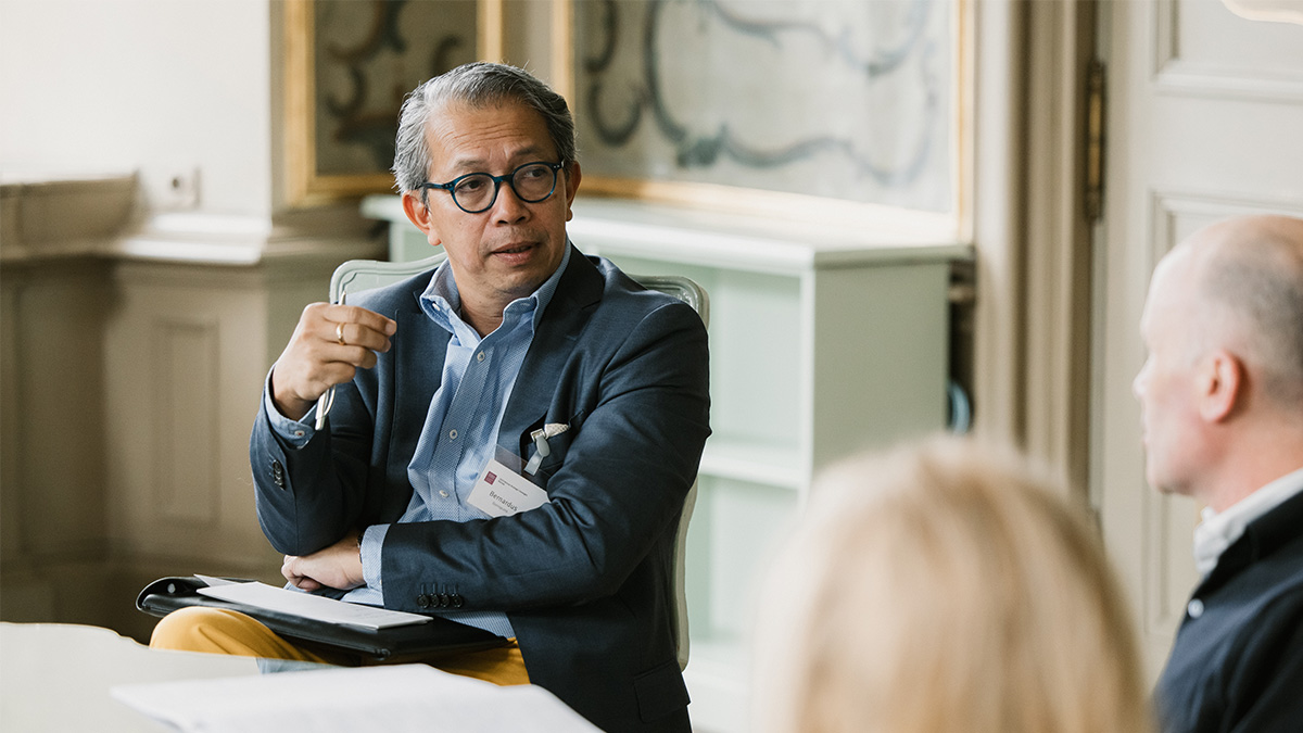 A man sits in a chair with his head turned to the right, talking to someone