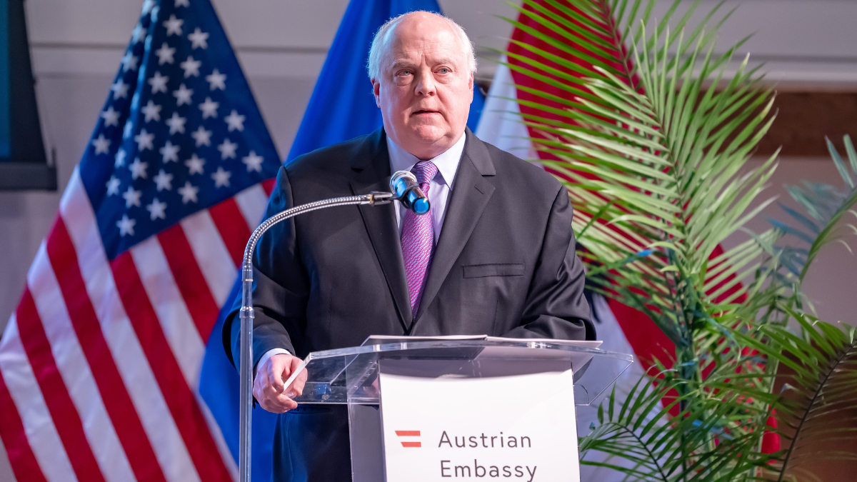 Hugh Verrier, a man with white hair, stands at a podium delivering a lecture in the Austrian Embassy in Washington. In the background are American and Austrian flags. 