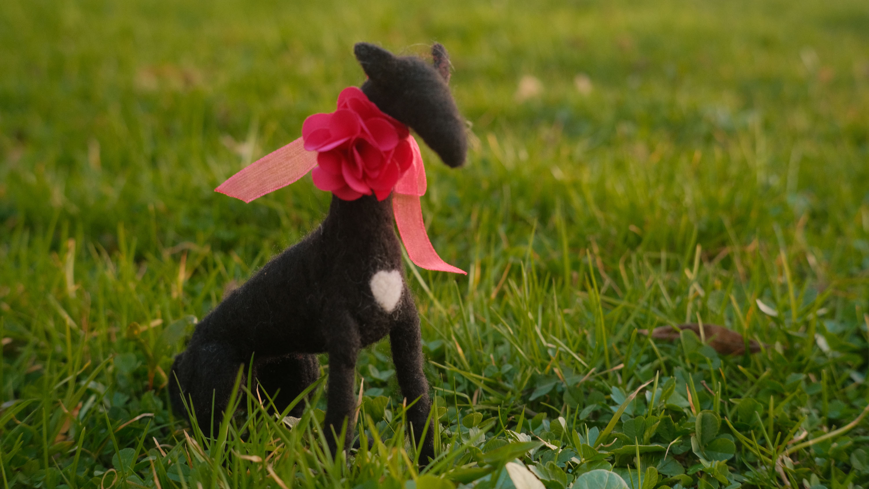 Needle felted symbol of Lucy Like-a-Charm, a black greyhound dog, wearing a pink ribbon, sitting in the grass, at the Schloss Leopoldskron.