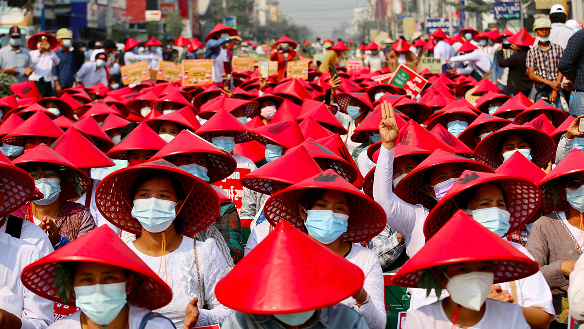 A large crowd of protesters wearing red conical hats and face masks gather peacefully, with some holding signs and making the three-finger salute.
